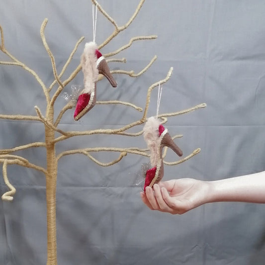 Showing the pair of burgundy velvet shoe hanging decorations on a twig tree with a model's hand holding the shoe by the tip