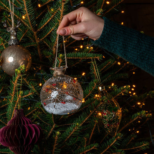 Light up glass bauble with snow and berries in hand in front of tree
