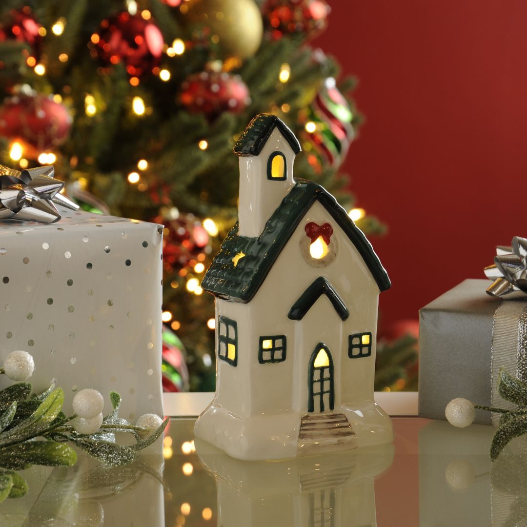 White light up house ornament with green roof and eaves and the lights on. Photographed in front of a Christmas tree and red wall