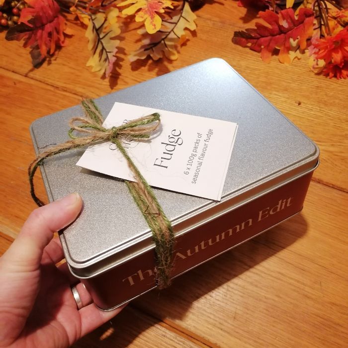 Fudge tin with a decorative bow on a wooden surface with autumn leaves.