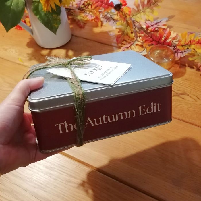 Hand holding a gift box labeled 'The Autumn Edit' on a wooden surface with autumn decorations.