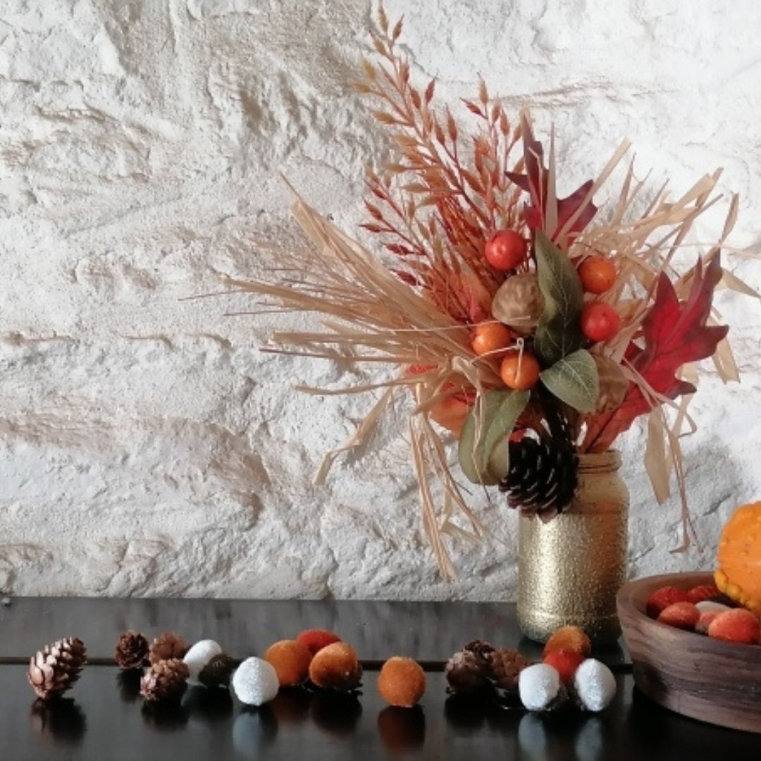 Autumn-themed floral arrangement with pumpkins and pinecones on a textured white wall background.