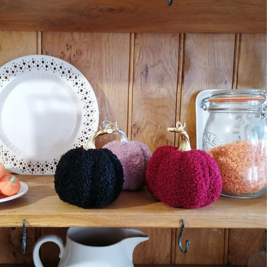 Three boucle wool pumpkins in black, pink, and red on a wooden surface with a jar and plate in the background.