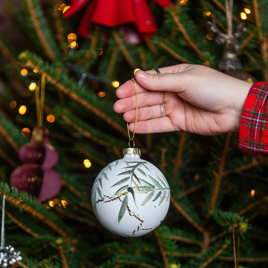 Hand holding a Christmas ornament with eucalyptus and marbling design in front of a decorated Christmas tree.