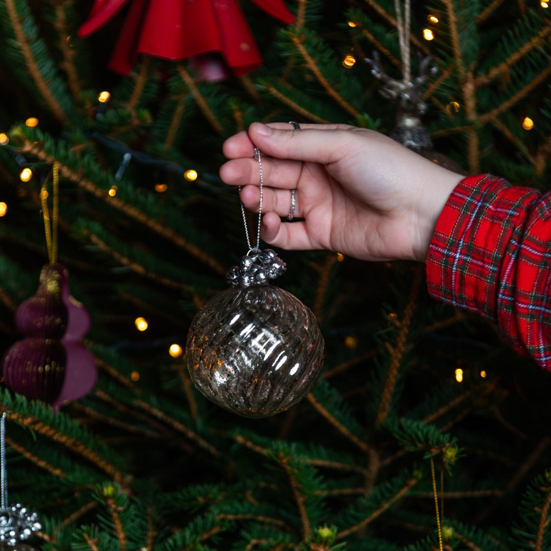 Hand holding a glass ornament in front of a decorated Christmas tree with lights.