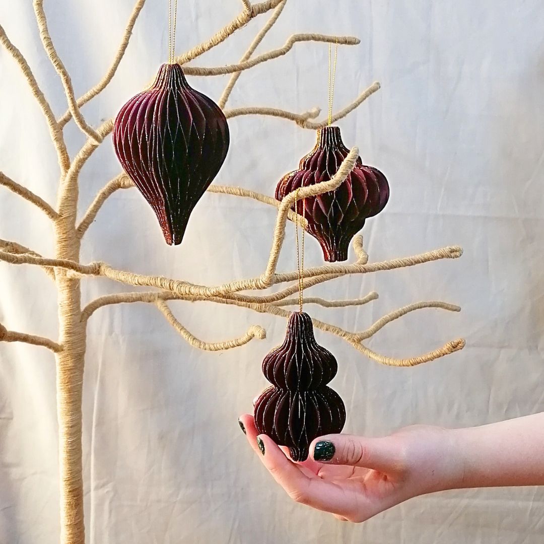 Showing the 3 different burgundy honeycomb paper decorations on a branch tree against a beige background, with a model's hand to show scale