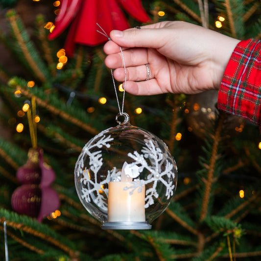 Lit up clear glass LED candle hanging bauble in hand in front of Christmas tree