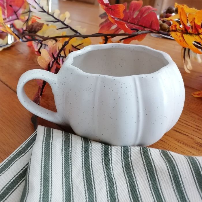White ceramic pumpkin-shaped mug on a wooden table with autumn leaves in the background.