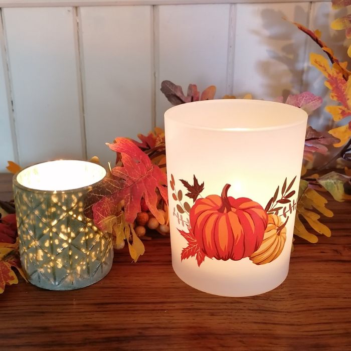 Two lit candles on a wooden surface with autumn leaves and pumpkins.