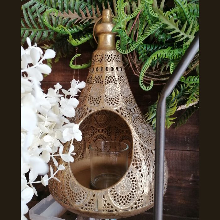 Decorative gold lantern with intricate patterns on a wooden surface with greenery and white flowers.