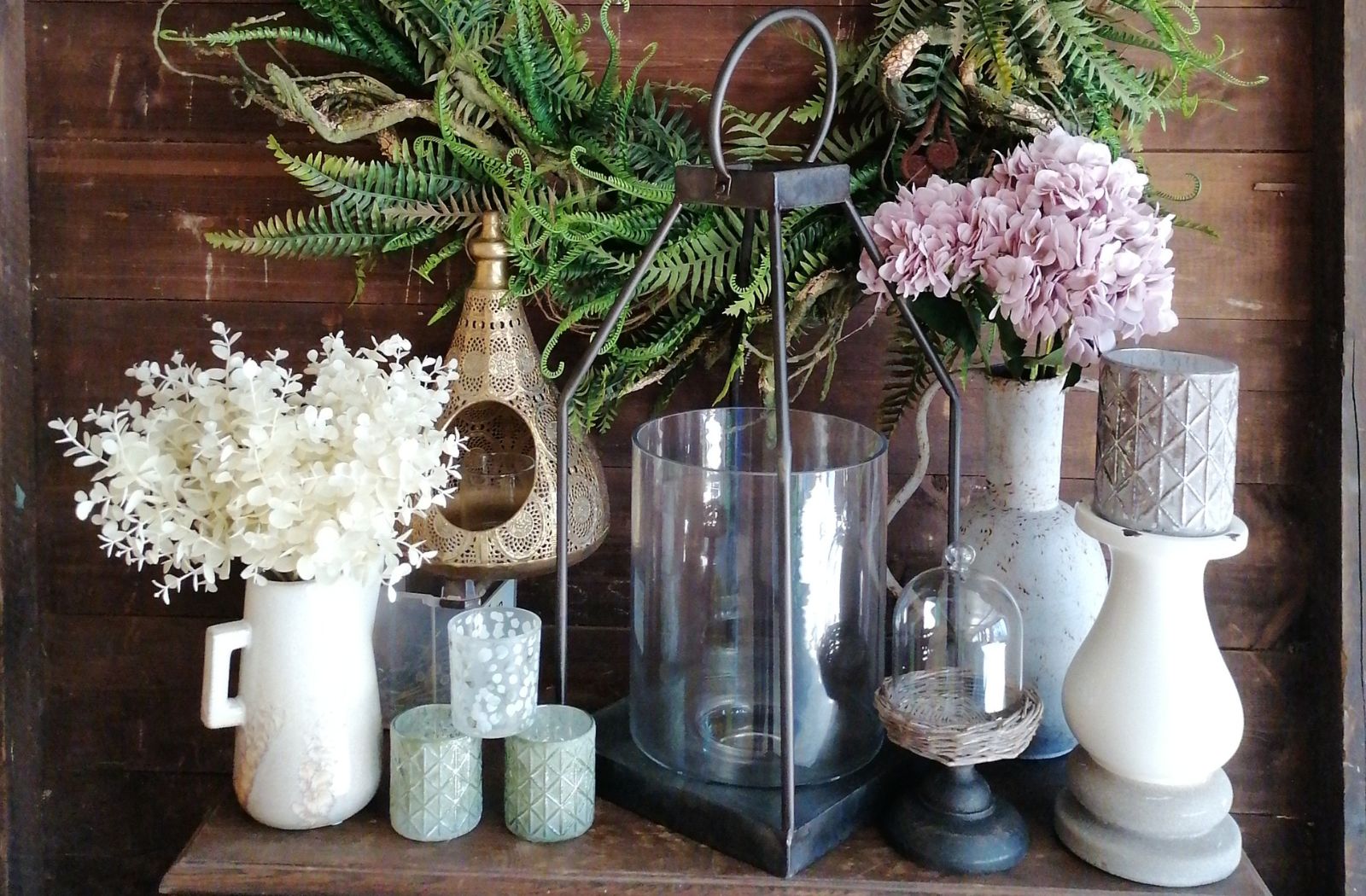 Decorative arrangement of vases, lanterns, and flowers on a wooden shelf against a wooden wall.