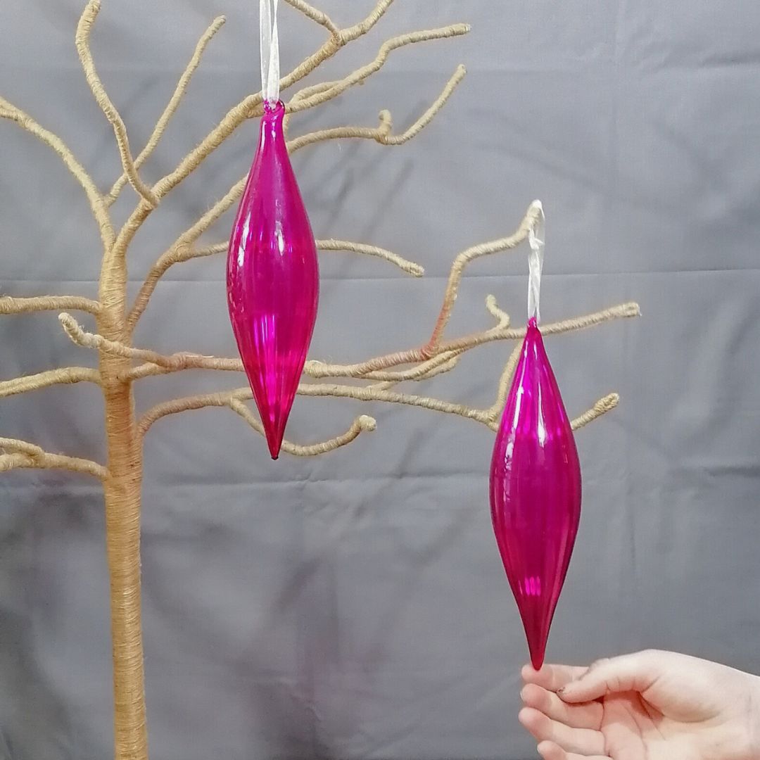 Showing the 2 hot pink glass hanging decorations on a branch tree with the model's hand to show scale