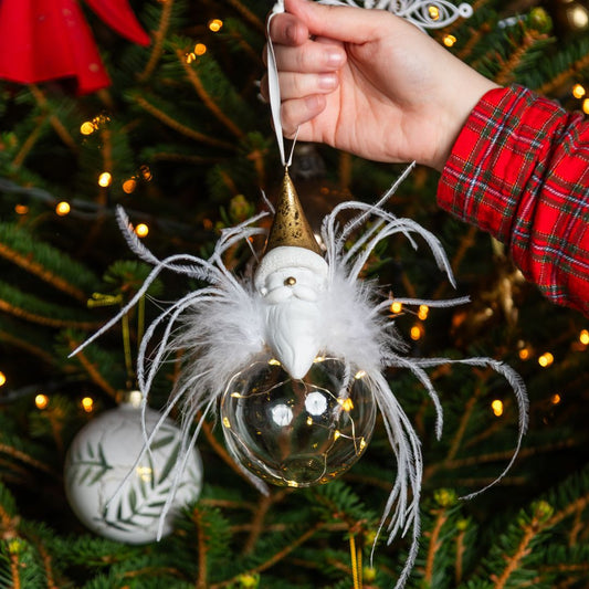 Decorative ornament with feathers and Father Christmas figure head held in hand against a Christmas tree