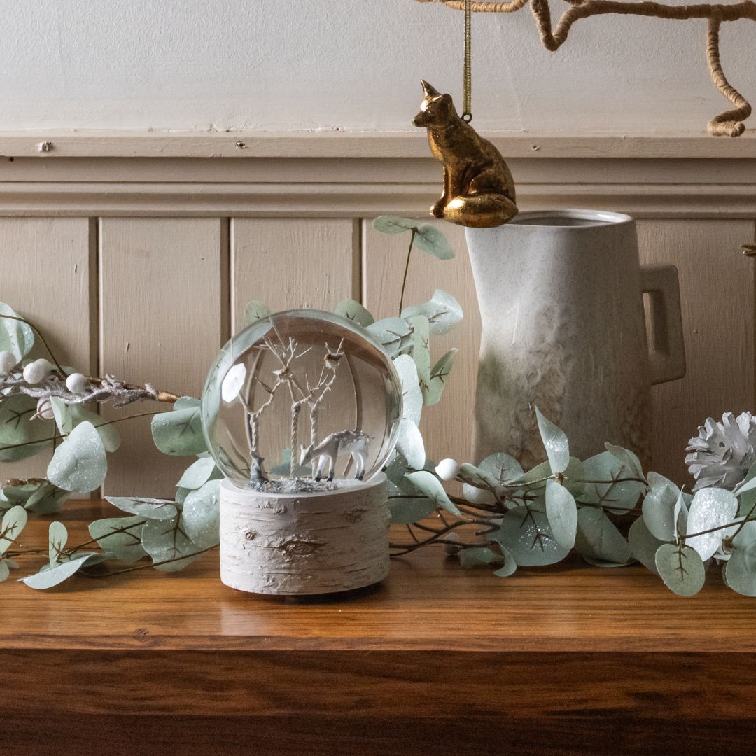 Decorative glass snowglobe with branches on a wooden surface with eucalyptus leaves and a ceramic jug.