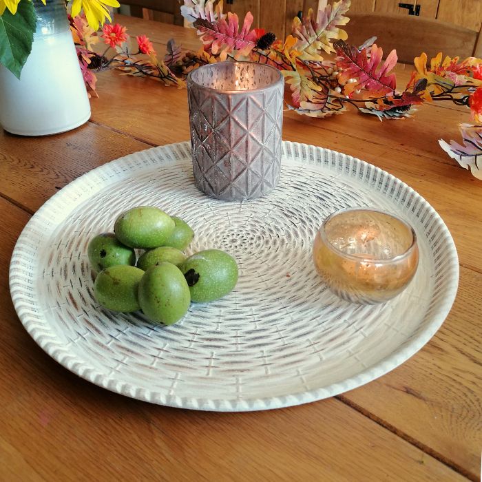 Decorative setting with a woven plate, green fruits, and candles on a wooden table.