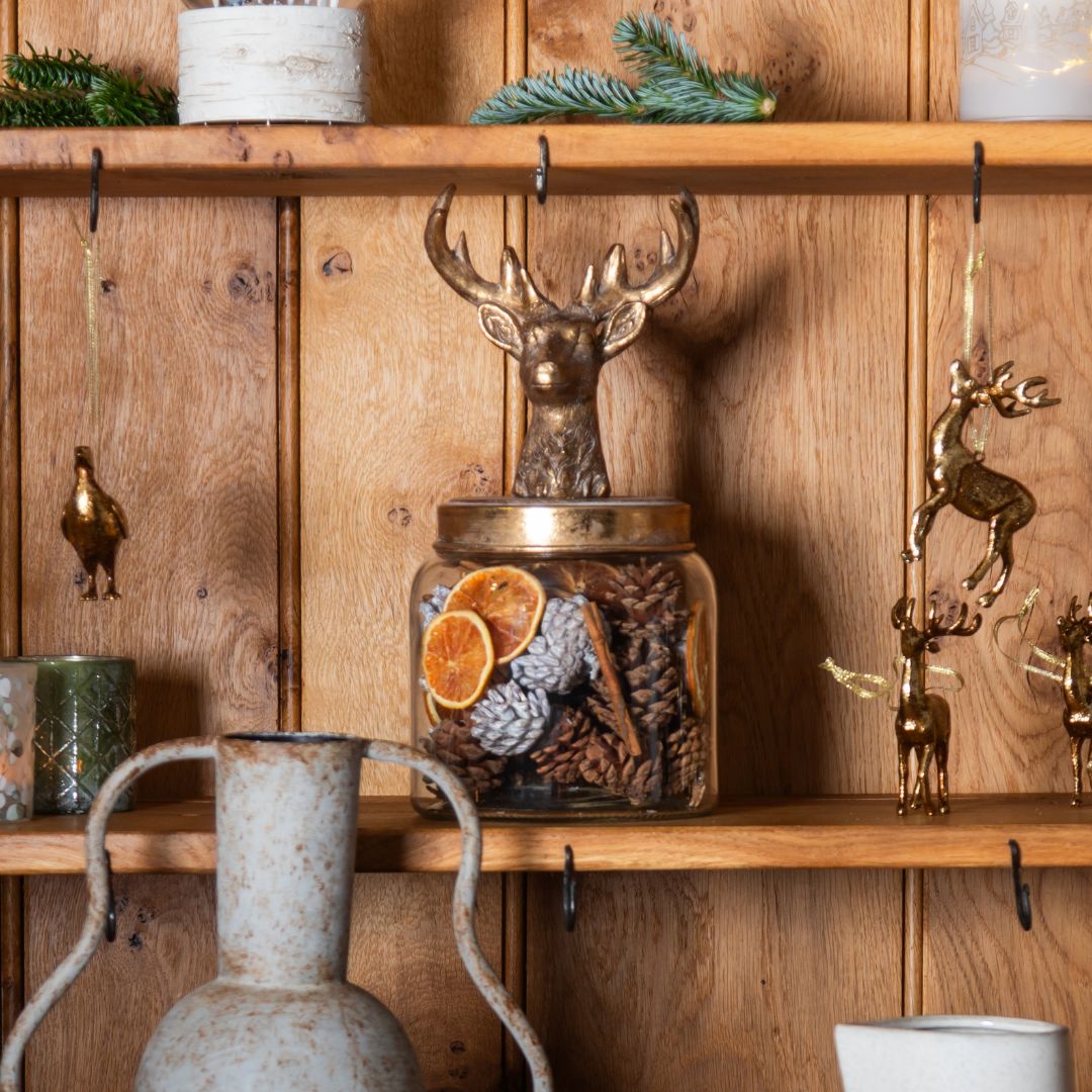 Decorative jar with dried oranges and pine cones on a wooden shelf with deer figurines.