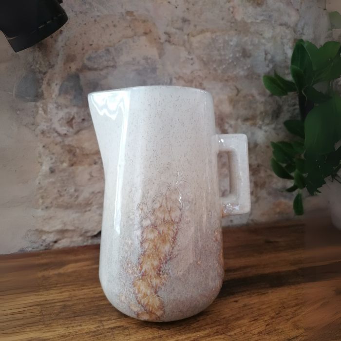 White ceramic pitcher with brown speckles on a wooden surface with a stone wall background