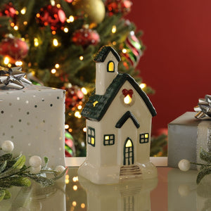 White light up house ornament with green roof and eaves and the lights on.  Photographed in front of a Christmas tree and red wall