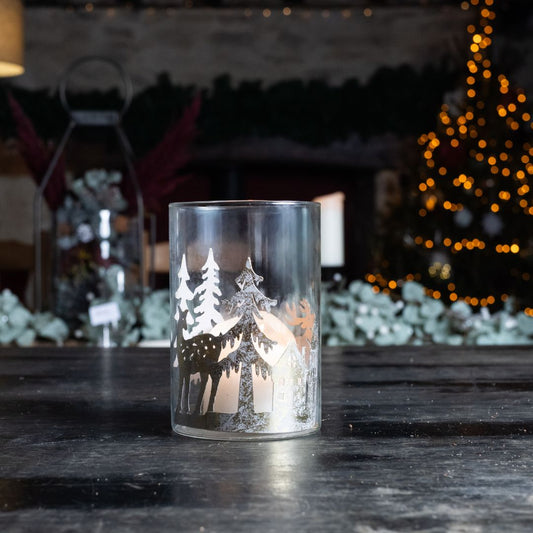 Decorative glass candle holder with winter scene on a dark surface, blurred festive background.