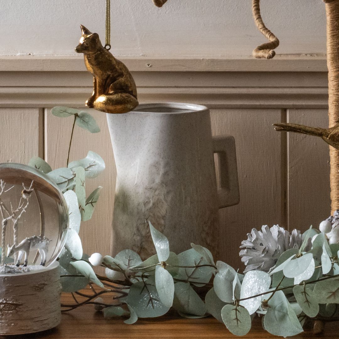 Decorative setup with a gold cat figurine, white jug, and greenery on a wooden surface.