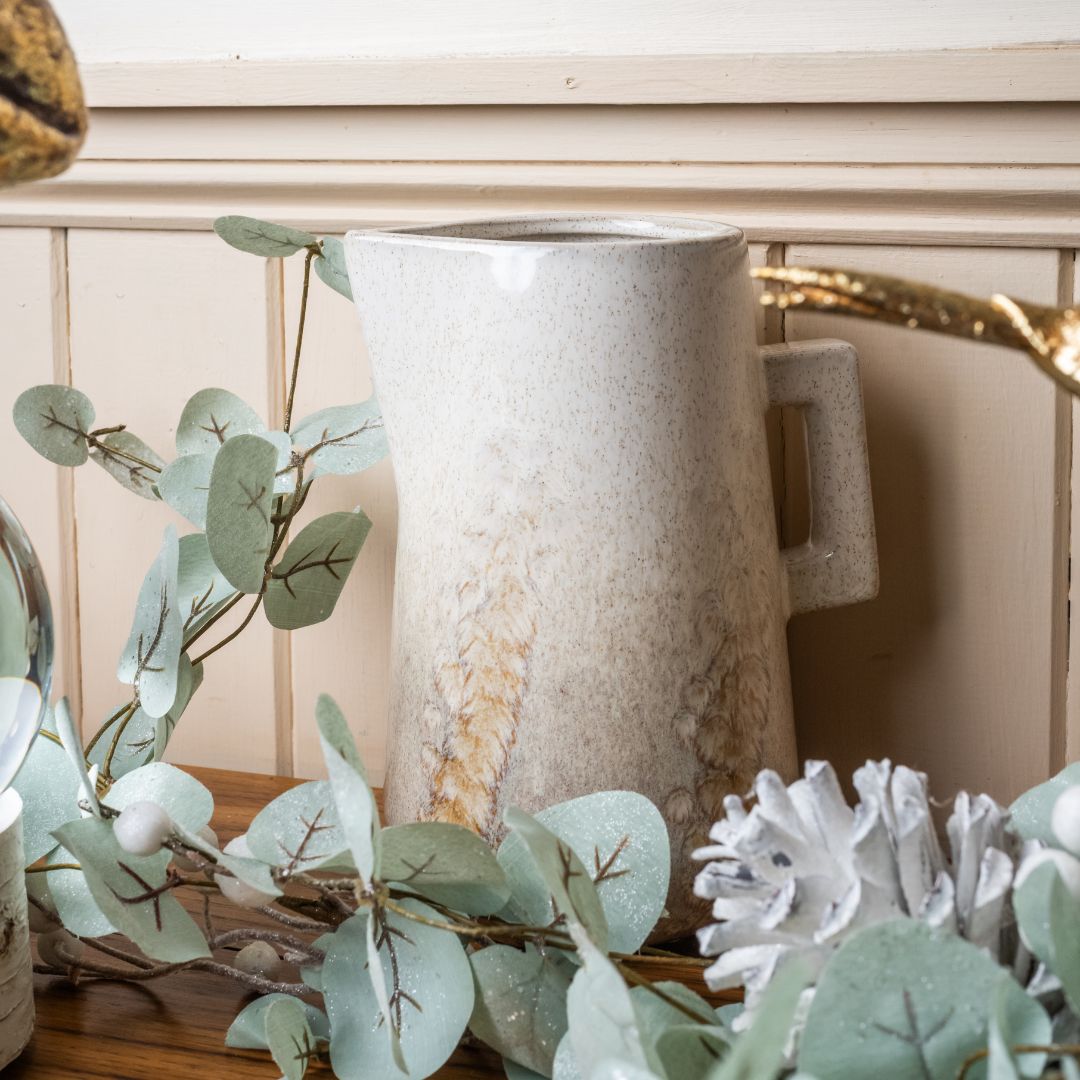 Decorative arrangement with a ceramic pitcher and greenery on a wooden surface.