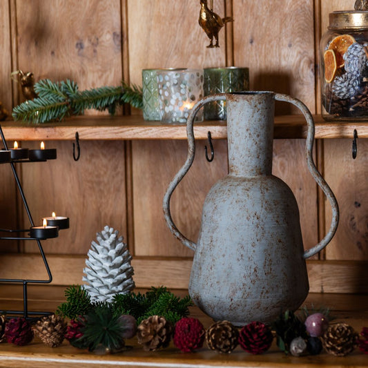 Decorative scene with a rustic vase, pine cones, and candles on a wooden shelf.