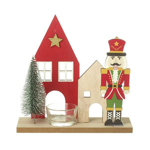 from left to right: a bristle tree with a red house behind it.  To the side of the red house a natural house that is smaller in height.  To the side of the natural house is a standing nutcracker with arms by his side.  The tealigth glass candle holder is in front fo the red house