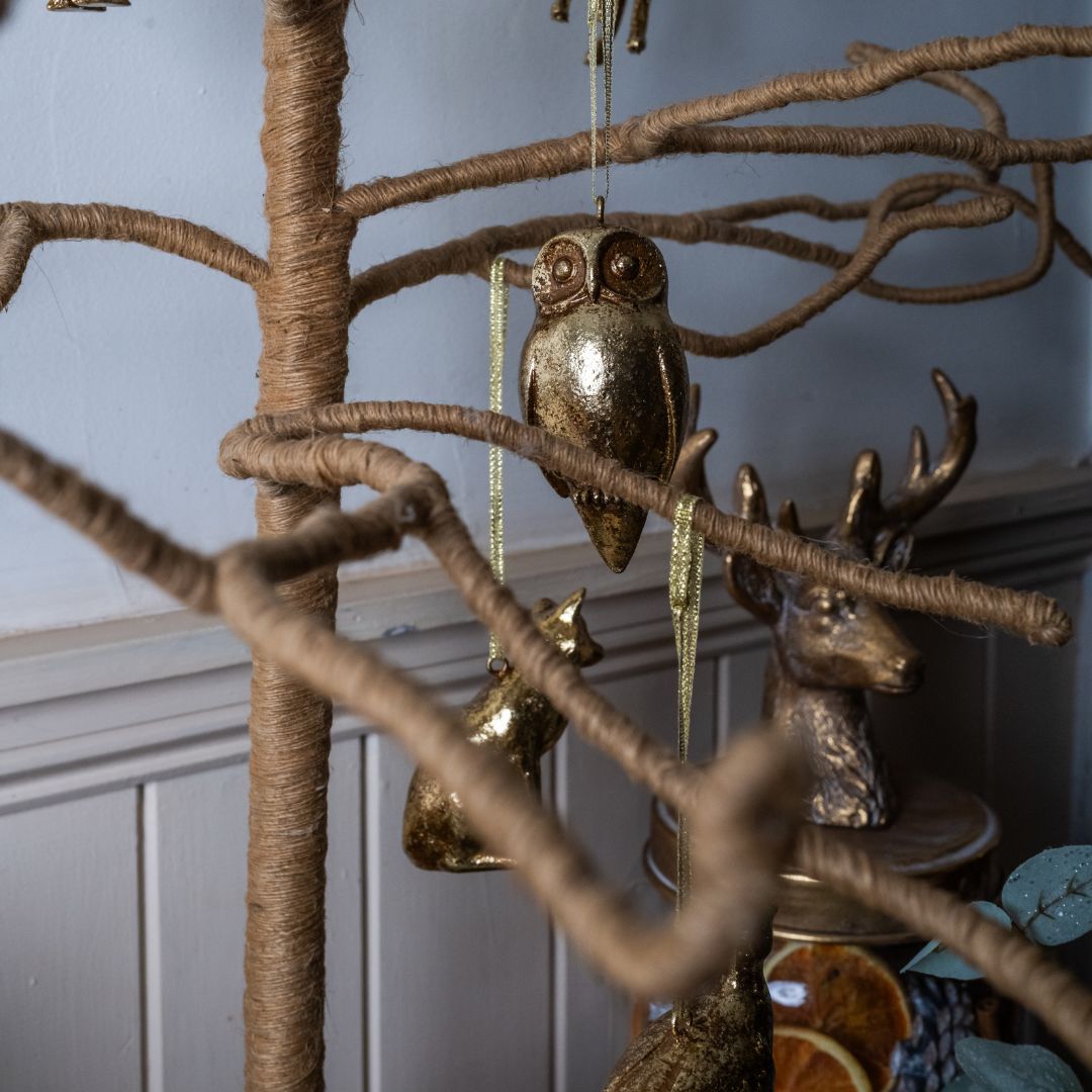 Decorative gold owl ornament hanging from a branch against a neutral wall.