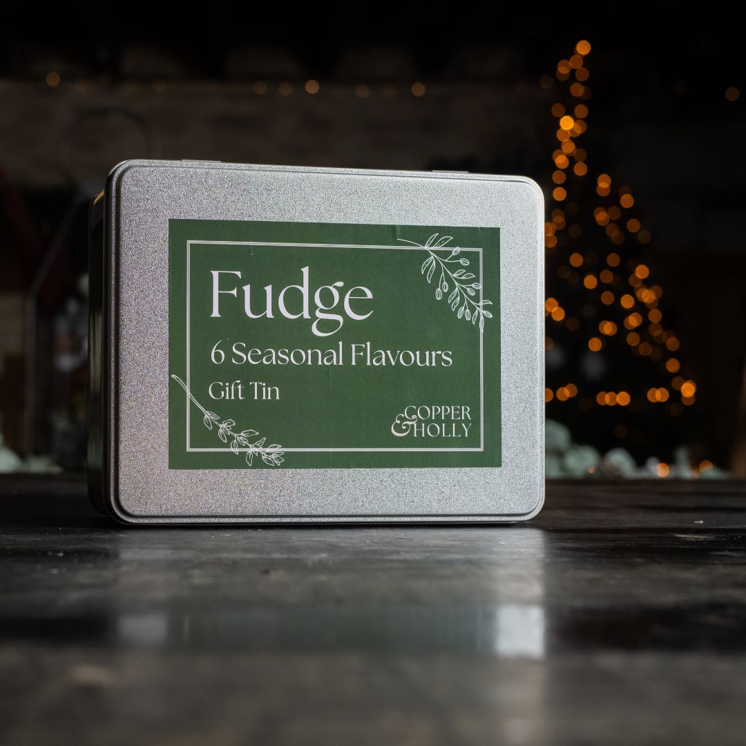 Fudge gift tin with 'Copper & Holly' branding on a dark surface with blurred lights in the background.