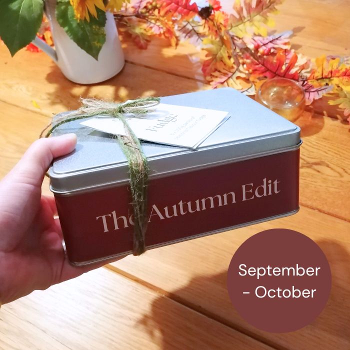 Hand holding a 'The Autumn Edit' seasonal fudge tin with fall-themed decor in the background.  Available September and October