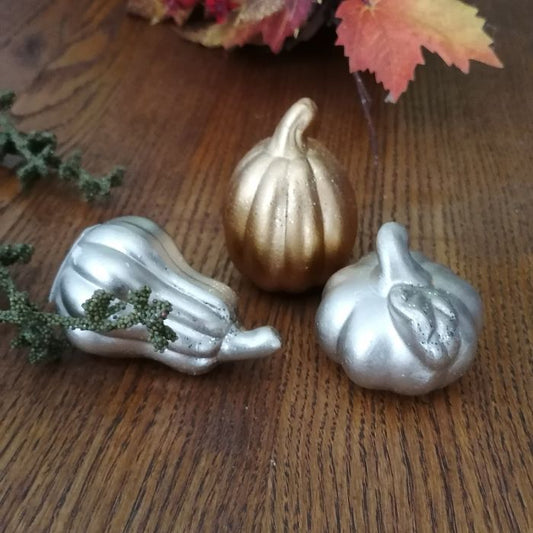 Three decorative pumpkins in silver and gold on a wooden surface with leaves.