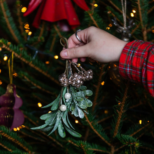 Hand holding mistletoe with gold berries in front of a decorated Christmas tree.