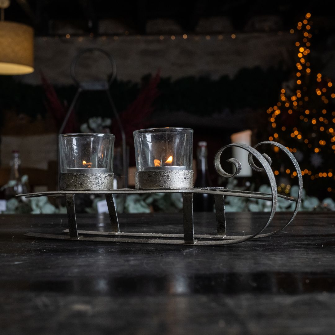 Two lit candles in glass holders on a metal sleigh with a blurred background