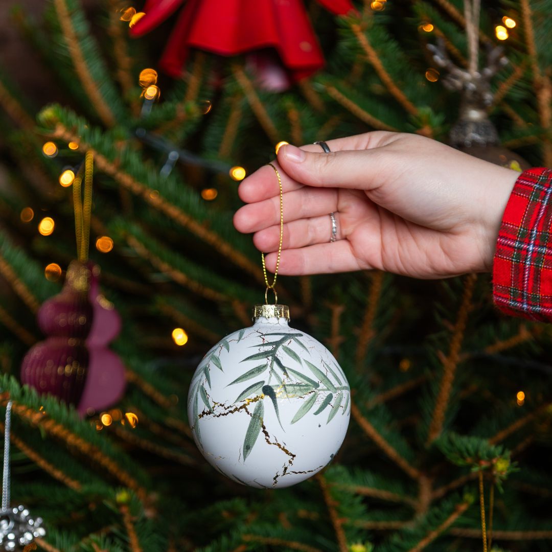 Hand holding a Christmas ornament with eucalyptus and marbling design in front of a decorated Christmas tree.