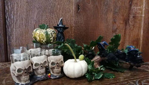 Set of 6 skull shaped shot glasses nestled in amongst oak leaves and with black cat decorations.  Shot against a dark oak wood background